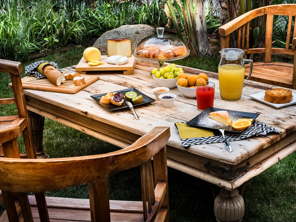 Teku Boutique Hotel a Bari Sardo – l’Ogliastra più autentica Colazione all’aperto su un tavolo di legno con formaggi, frutta, pane e succhi