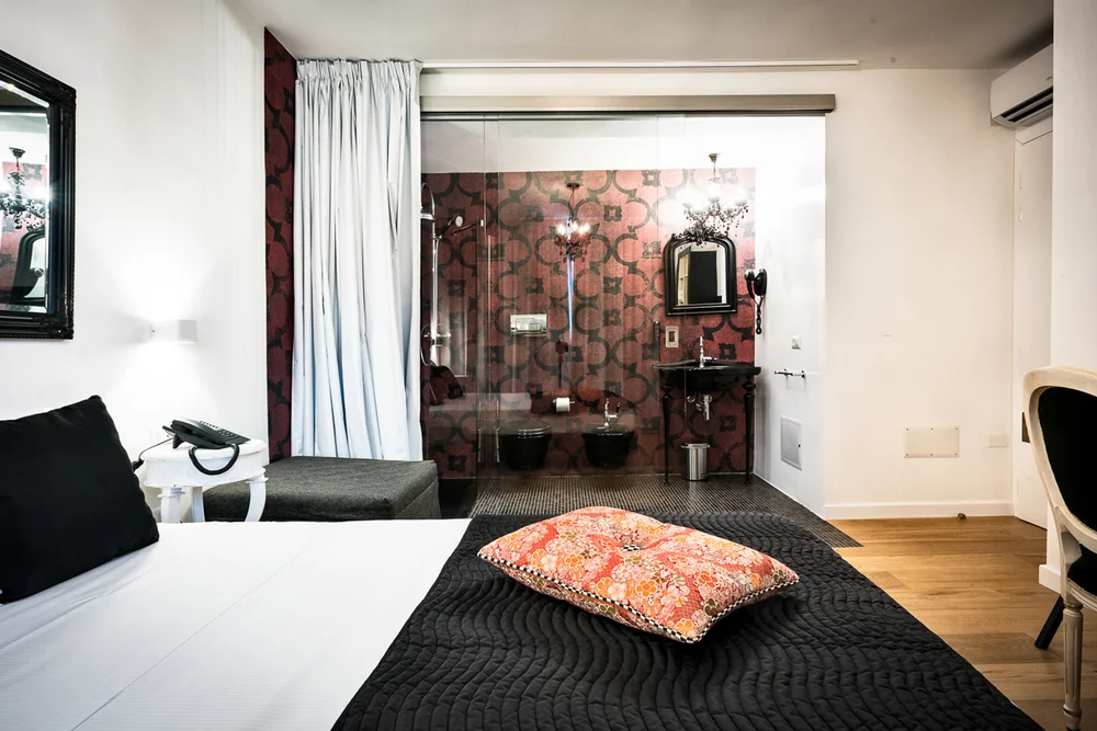 Camera d’hotel con letto, coperta nera e cuscino floreale, bagno a vista dietro una parete di vetro
