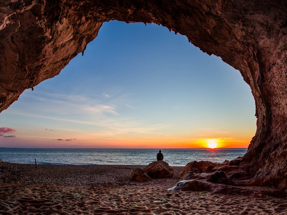 Teku Boutique Hotel a Bari Sardo – l’Ogliastra più autentica Una persona osserva il tramonto sul mare dall’interno di una grotta