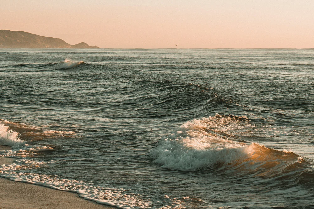 Wellen brechen bei Sonnenuntergang an einem Sandstrand, in der Ferne ein Küstenvorsprung