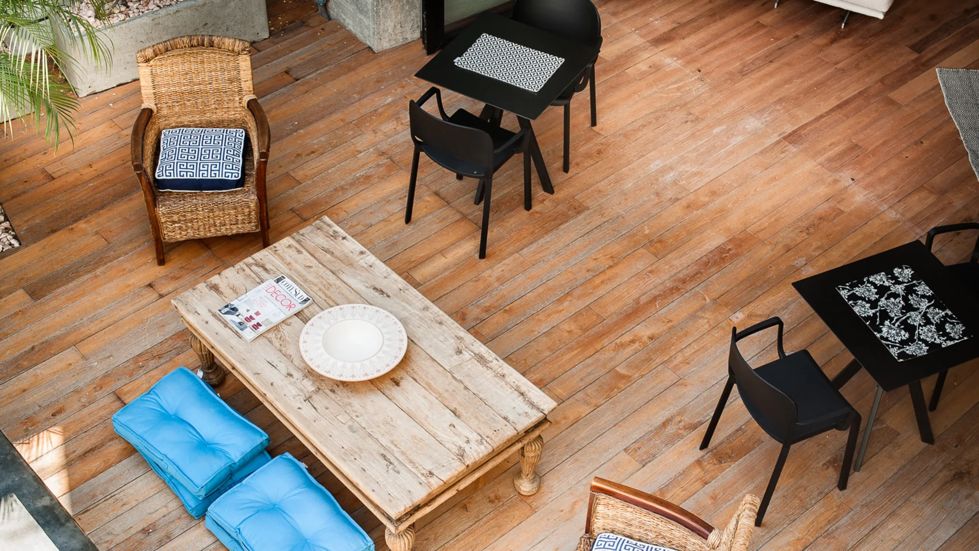 Terrazza in legno con sedie in vimini, tavolini neri, cuscini blu e piante
