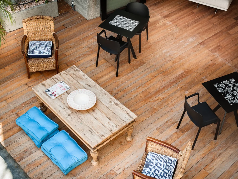 Teku Boutique Hotel a Bari Sardo – l’Ogliastra più autentica Terrazza in legno con sedie in vimini, tavolini neri, cuscini blu e piante