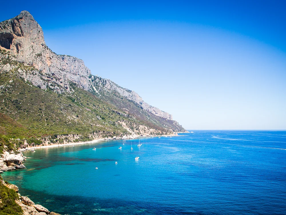 Teku Boutique Hotel a Bari Sardo – l’Ogliastra più autentica Scogliere rocciose sopra una baia turchese con piccole barche e cielo sereno