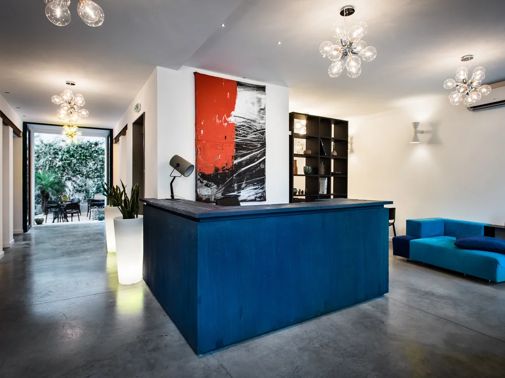 Teku Boutique Hotel a Bari Sardo – l’Ogliastra più autentica Reception moderna con bancone blu, divano turchese e accesso a un giardino
